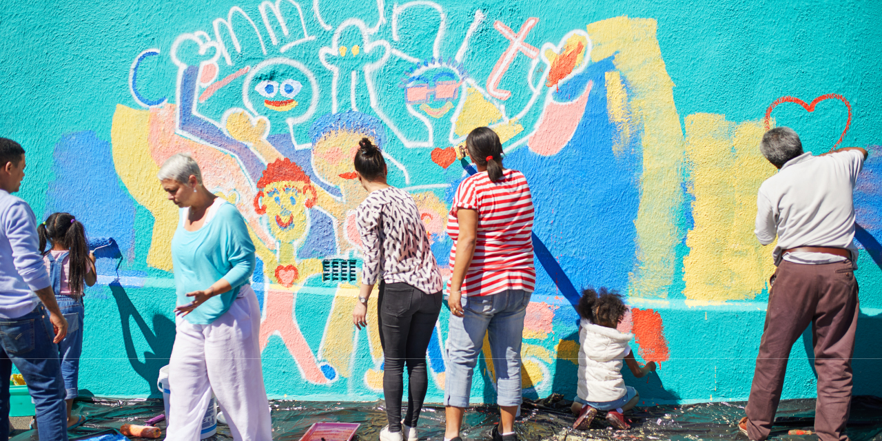  Five adults, two kids paint a colorful mural of the word ‘community’ over smiling people on a building’s outside wall