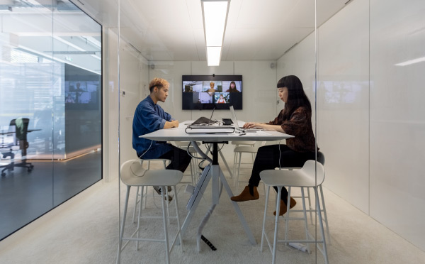 An image of a man and woman attending a hybrid meeting inside a glassed-in conference room. 
