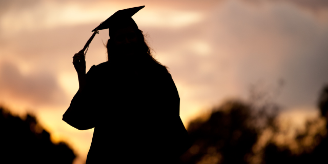A graduate in cap and gown is silhouetted by sunset.