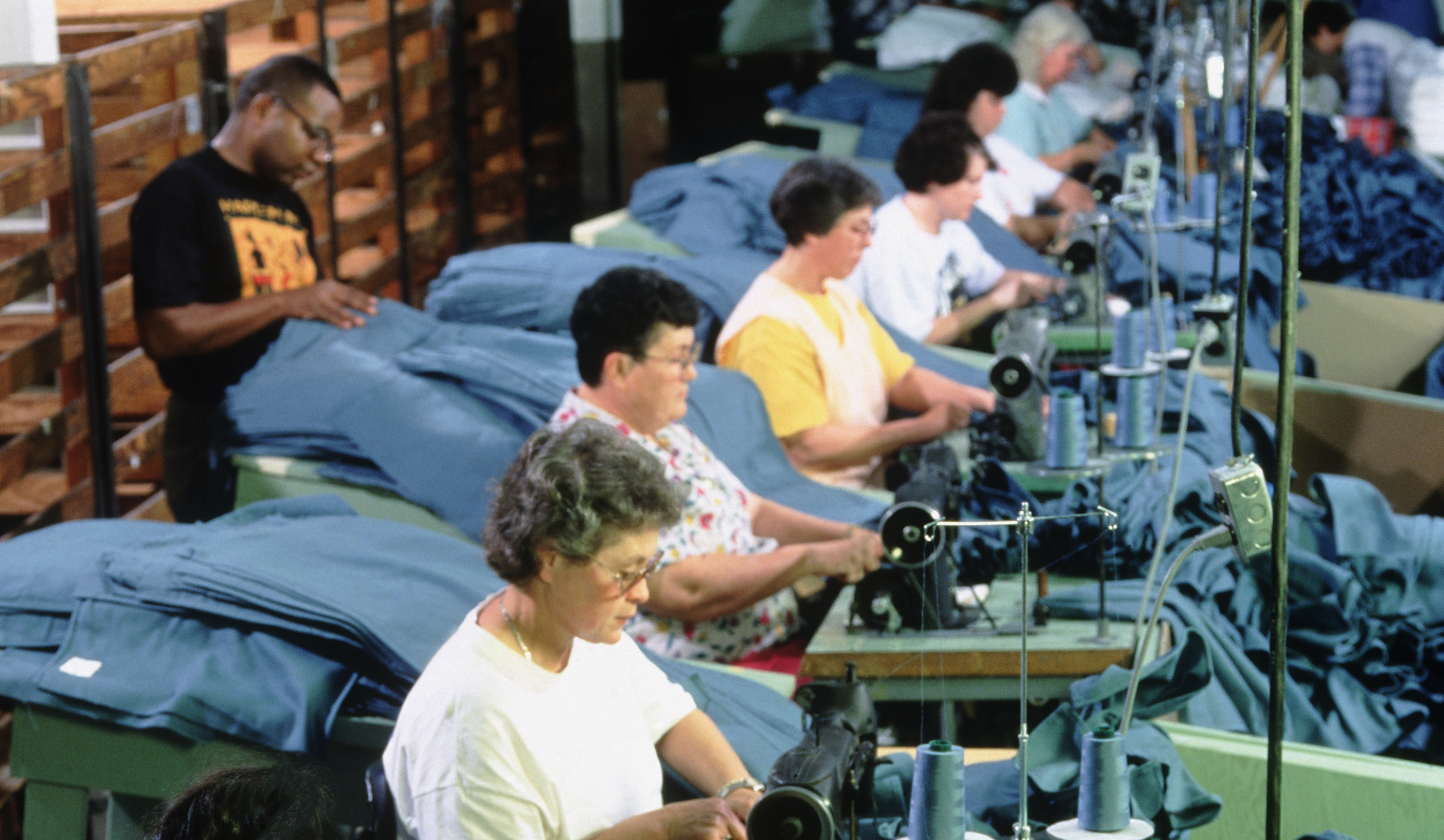 Photo of men and women sitting at sewing machines all in a row handling blue material.