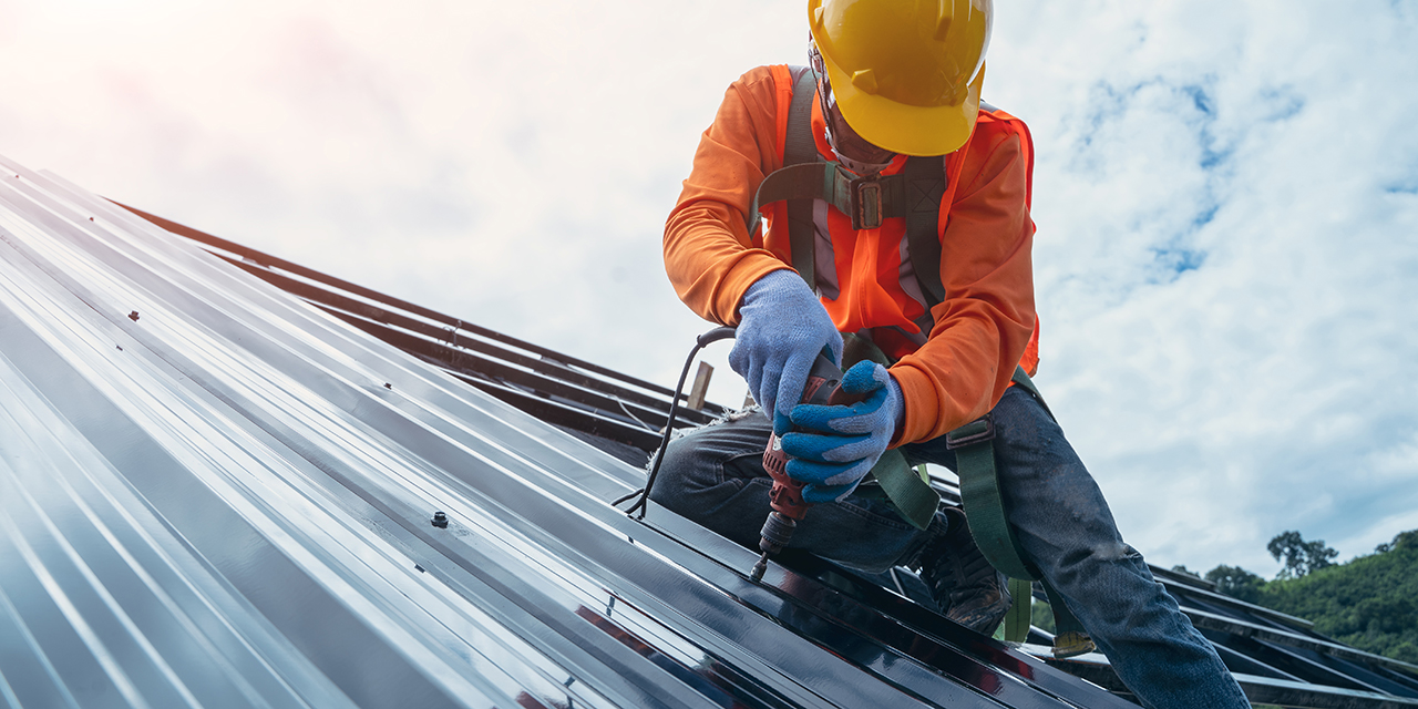 professional roofer in orange jacket and yellow hardhat installing metal roofing panels against a cloudy sky