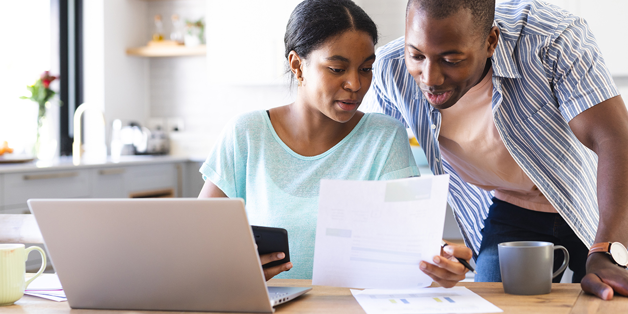 Woman sitting at kitchen table with daughter on lap looking at a bill with laptop open in front of them