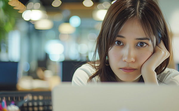 AI image of young brunette woman sitting in an office and staring at her computer, looking bored. One elbow is on the desk with her arm up toward her face and a hand cupping her cheek.