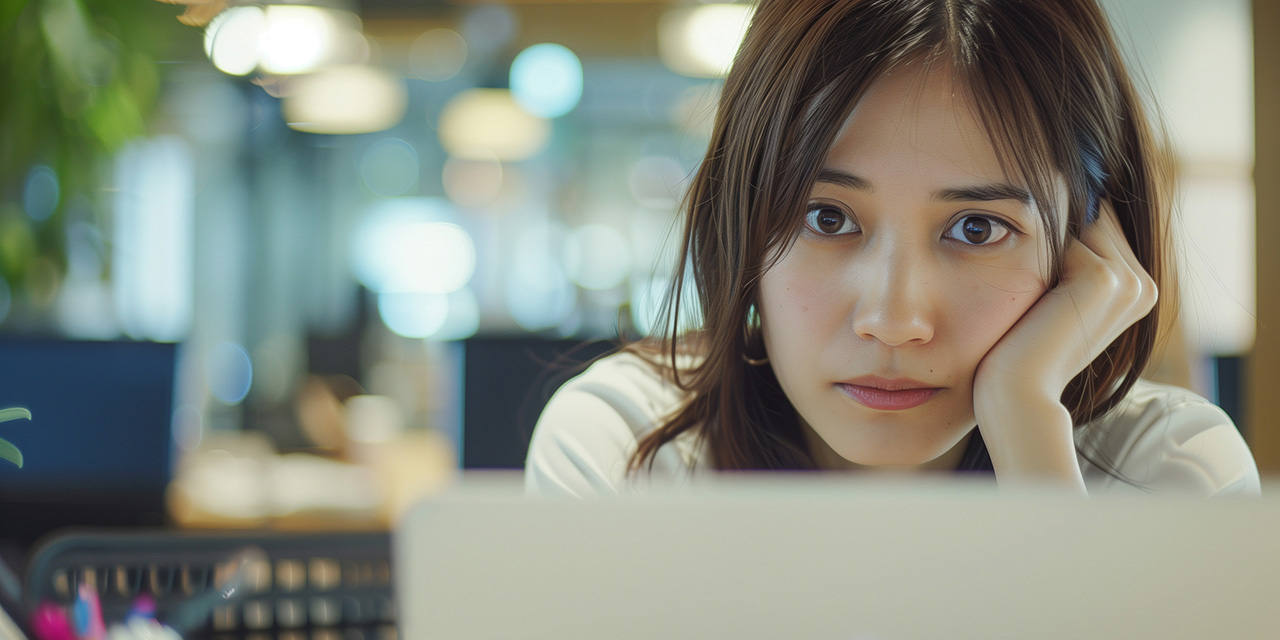 AI image of young brunette woman sitting in an office and staring at her computer, looking bored. One elbow is on the desk with her arm up toward her face and a hand cupping her cheek.